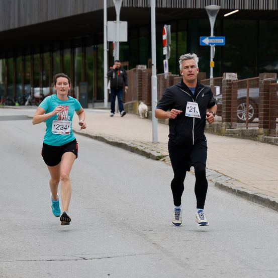 Zwei Läufer nehmen an einem Marathon teil. Eine Frau mit der Nummer 234 und ein Mann mit der Nummer 21. Sie laufen auf einer asphaltierten Straße mit einem Ziegelsteinpflaster. Hinter ihnen geht ein Mann mit einem Hund.