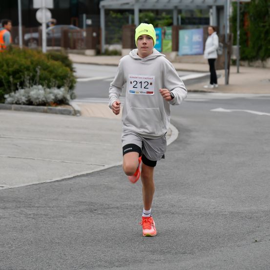 Ein Läufer mit der Nummer 212 auf dem Shirt joggt auf einer Straße. Im Hintergrund steht eine Person in der Nähe eines Gebäudes.