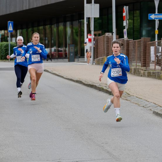 Drei Läuferinnen nehmen an einem Marathon auf einer Straße teil. Zwei sind vorne, eine dahinter. Sie tragen passende blaue Trikots mit Nummern.