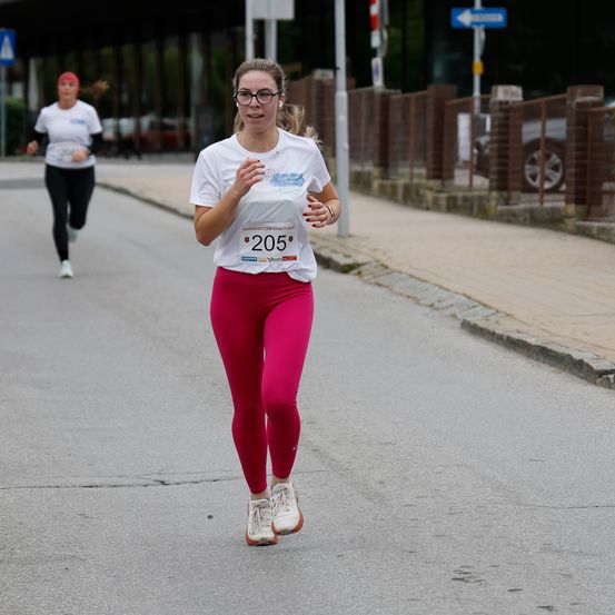 Eine Frau mit Brille und einem weißen Shirt mit der Nummer 205 läuft auf einer Straße beim Marathon. Eine andere Frau läuft hinter ihr.