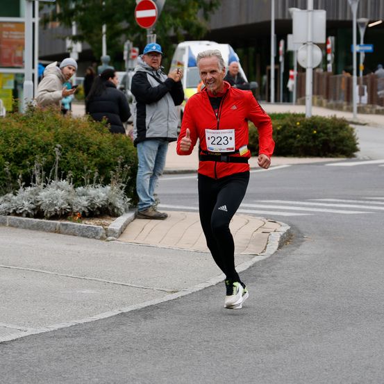 Ein älterer Mann in einem roten Jackett mit der Startnummer 223 läuft einen Marathon. Zuschauer beobachten ihn von der Straßenseite.