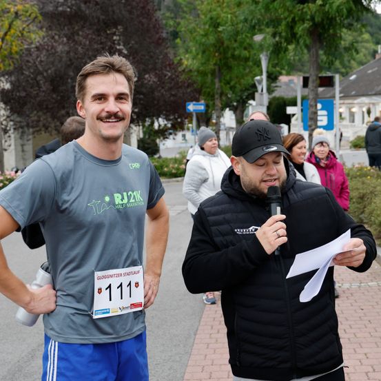 Ein Mann in einem Marathon-T-Shirt steht draußen. Ein anderer Mann hält ein Mikrofon und Papier. Dahinter laufen mehrere Menschen. Bäume und Gebäude sind im Hintergrund.