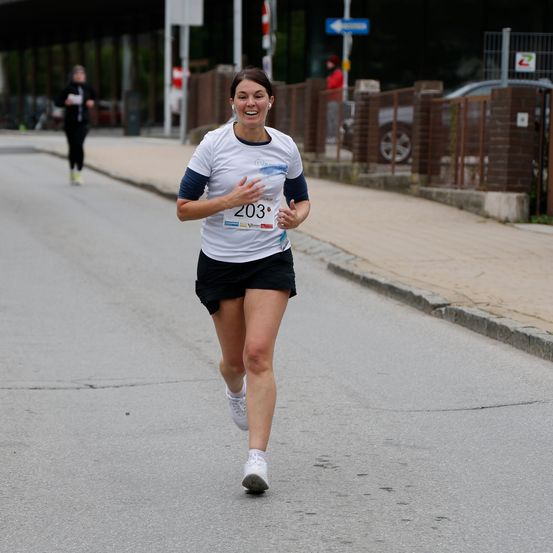 Eine Frau in einem weißen T-Shirt mit der Nummer 203 auf der Brust läuft auf der Straße. Eine andere Person geht hinter ihr. Der Bürgersteig hat einen Metallzaun und eine Ziegelmauer. Vor dem Gebäude sind Autos geparkt.