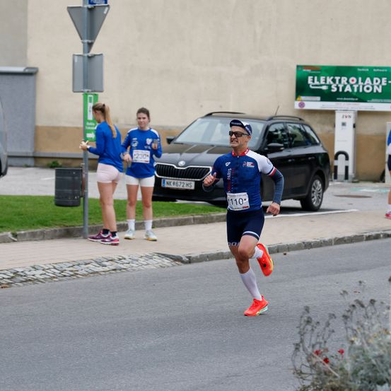 Ein Läufer mit Sonnenbrille und einer Nummer 110 auf dem Trikot läuft eine Straße hinunter, während zwei Frauen in blauen Trikots auf dem Bürgersteig stehen. Ein schwarzer SUV mit dem Kennzeichen NK 672 HRS ist geparkt. Eine Elektroladestation ist rechts zu sehen.