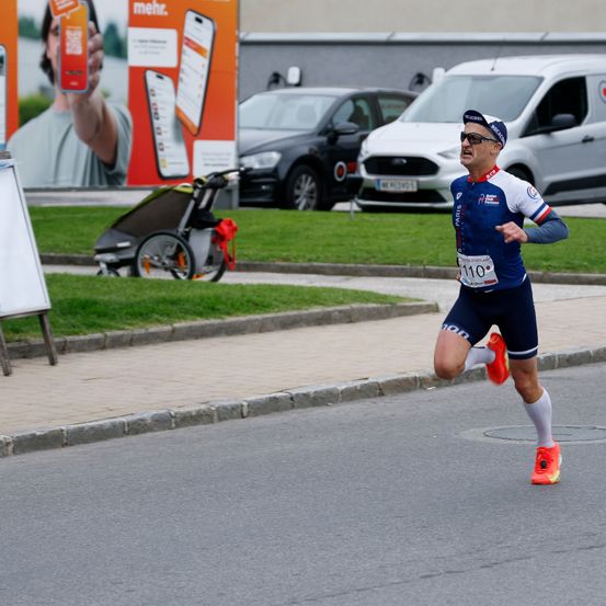 Ein Mann läuft bei einem Marathon in einem blau-weißen Outfit mit der Nummer 110 auf den Shorts. Es gibt zwei Fahrzeuge und einen Kinderwagen an der Seite.