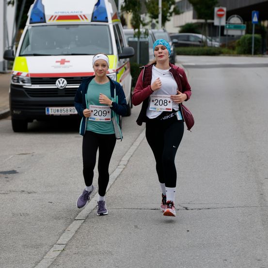 Zwei Frauen laufen auf der Straße. Sie tragen Nummern auf ihren Shirts, 208 und 209. Ein Van ist hinter ihnen geparkt.