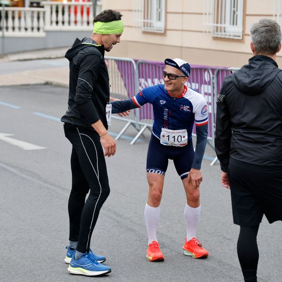 Ein Mann in einem Radsport-Outfit mit der Startnummer 110 unterhält sich mit einem lächelnden Mann in schwarzer Sportbekleidung, während eine weitere Person zusieht.