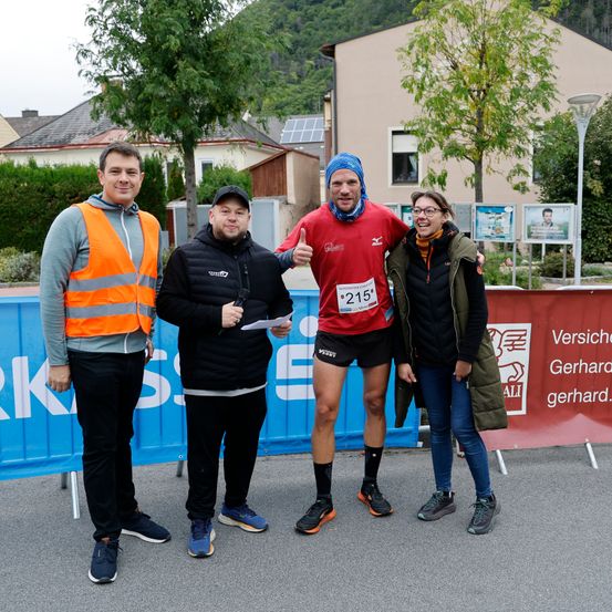 Vier Personen stehen vor einem blauen Banner mit weißen Buchstaben. Der Mann in der Mitte trägt ein rotes Shirt mit der Nummer 215. Die anderen tragen Jacken und Sneaker. Dahinter sind Häuser, Bäume und ein rotes Banner mit weißen Buchstaben.