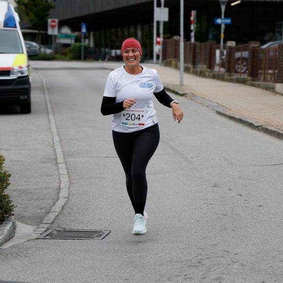 Eine Frau in einem weißen T-Shirt mit der Nummer 204 darauf läuft mitten auf der Straße. Sie lächelt und scheint an einem Marathon teilzunehmen.