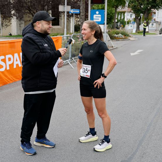Ein Läufer beim Marathon wird von einem Mann mit Mikrofon interviewt. Der Läufer trägt ein schwarzes Shirt mit der Nummer 35 und Laufshorts.