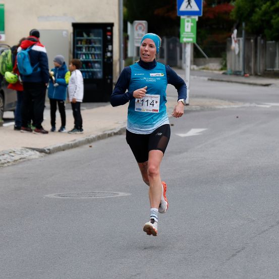 Eine Frau mit einem blauen Stirnband läuft bei einem Marathon auf einer asphaltierten Straße. Zuschauer stehen hinter ihr auf dem Bürgersteig.