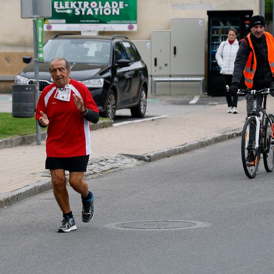 Ein älterer Mann joggt auf einer Straße, neben ihm fährt ein Fahrradfahrer und eine Frau geht auf dem Bürgersteig.