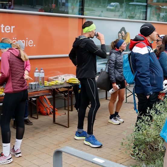 Eine Gruppe von Menschen, wahrscheinlich Athleten, ist versammelt. Ein Mann trinkt aus einer Flasche. Bananen und Wasserflaschen stehen auf einem Tisch. Eine Frau trägt einen Rucksack.
