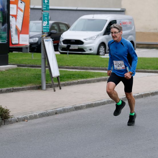Ein älterer Mann läuft auf einer asphaltierten Straße und trägt ein blaues Shirt und schwarze Shorts mit der Nummer 126. Dahinter befindet sich ein weißer Lieferwagen und ein schwarzes Auto.