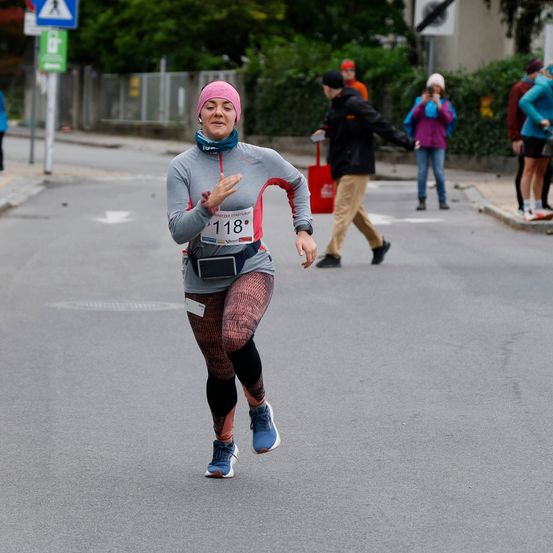 Eine Läuferin, die eine pinke Mütze und ein graues Sweatshirt trägt, läuft in einem Marathon. Sie trägt die Startnummer 118. Dahinter laufen mehrere Menschen auf dem Bürgersteig.
