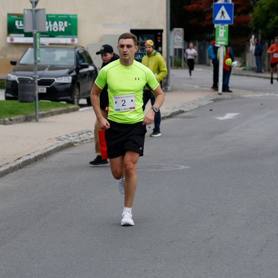 Ein Mann in einem grünen Shirt und schwarzen Shorts läuft einen Marathon auf einer Straße. Im Hintergrund ist ein Auto geparkt.