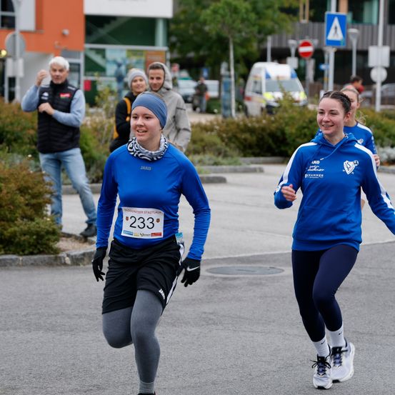 Zwei Frauen laufen bei einem Marathon, eine mit der Nummer 233. Ein Mann jubelt von der Seite. Ein weißer Van ist in der Nähe geparkt. Bäume und ein Gebäude im Hintergrund.
