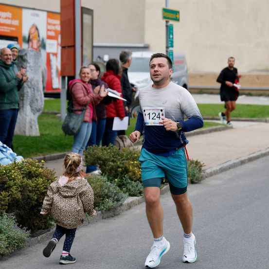 Ein Mann mit einem Startnummernband läuft bei einem Marathon. Zuschauer beobachten von den Seitenlinien, und ein kleines Kind folgt ihm dicht auf den Fersen.