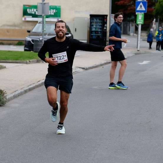 Ein Mann in einem schwarzen Shirt und Shorts läuft auf einer Straße mit der Nummer 122 auf der Brust. Eine weitere Person geht hinter ihm auf dem Bürgersteig.