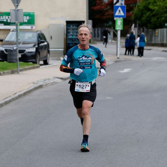 Ein Mann läuft auf einer Stadtstraße einen Marathon. Er trägt ein blaues Shirt mit der Nummer 127. Hinter ihm steht ein schwarzer Wagen auf dem Rasen.