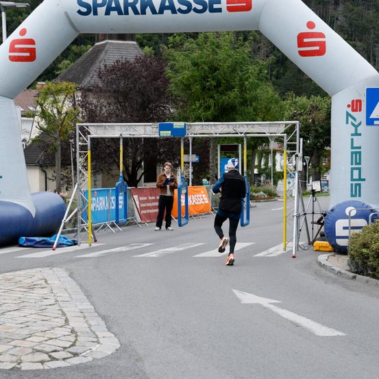 Ein Läufer mit Helm überquert die Ziellinie eines Rennens unter einem Sponsorenbanner. Hinter ihm steht eine Frau an einem Stand mit Mikrofon.