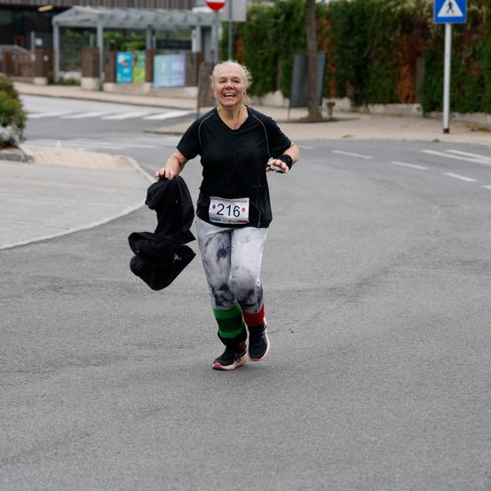 Eine Frau lächelt und läuft einen Marathon mit der Nummer 216 auf ihrem Shirt. Sie hält eine Jacke in ihrer linken Hand.