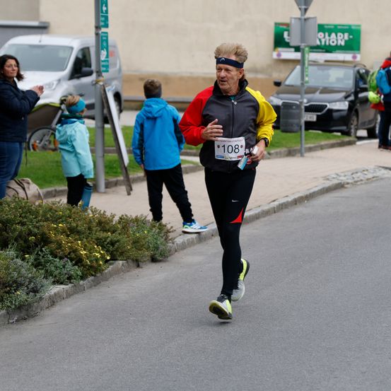 Ein älterer Mann läuft einen Marathon mit der Nummer 108 auf seinem Trikot. Hinter ihm beobachten zwei Kinder und eine Frau. Am Straßenrand sind Autos und ein Van geparkt.