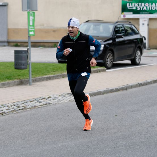 Ein Mann in Sportbekleidung läuft auf einer Straße, in der Nähe ist ein schwarzer SUV geparkt. Ein Schild befindet sich auf der linken Seite und ein Gebäude mit einem grünen Banner im Hintergrund.