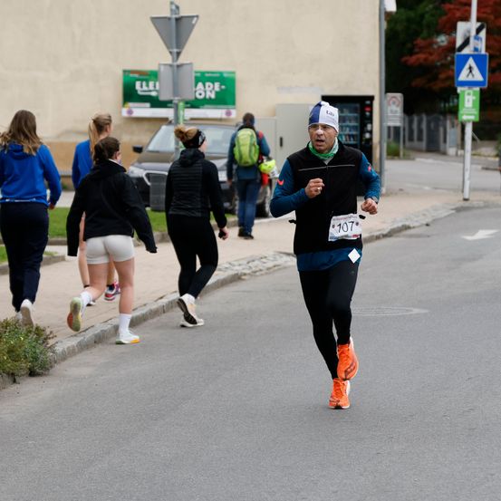 Ein Mann läuft in einem Marathon mit einer weißen Mütze, einer schwarzen Jacke und orangefarbenen Schuhen. Auf seiner Weste ist die Nummer 107 zu sehen. Hinter ihm gehen Menschen auf dem Bürgersteig. Im Hintergrund befinden sich Gebäude, Autos und Bäume.