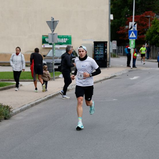 Ein Mann läuft auf einer Straße mit der Nummer 8 auf seinem Shirt. Mehrere Leute gehen auf dem Bürgersteig, und einige tragen Taschen. Es gibt Bäume, Straßenschilder und einen Verkaufsautomaten am Straßenrand.