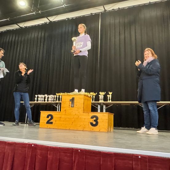 Ein Mädchen steht auf dem Podest des ersten Platzes und hält eine Trophäe, während zwei Frauen applaudieren. Die Bühne hat ein Holzpodest mit den Nummern 1, 2 und 3. Hinter dem Podest sind Trophäen auf einem Tisch angeordnet.
