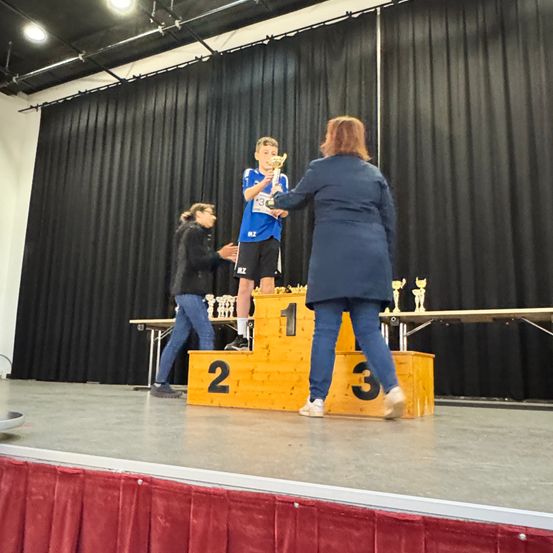 Ein junger Junge steht auf dem Siegerpodium, einen Pokal in der Hand, neben zwei Frauen. Dahinter steht ein Tisch mit Pokalen.