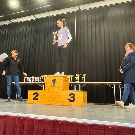Ein Mädchen steht auf einem Podium mit der Nummer 1 und hält einen Pokal. Zwei Frauen sind mit ihr auf der Bühne, eine hält ein Stück Papier. Der Podest ist von Pokalen umgeben. Hinter dem Podium steht ein Tisch mit Pokalen darauf und ein schwarzer Vorhang.
