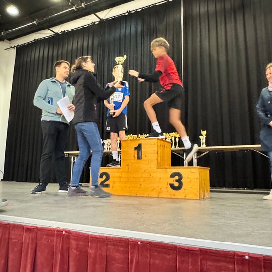 Ein junger Athlet steht auf einem Podest mit der Nummer 1 und hält einen Pokal, während zwei Erwachsene ihn präsentieren. Dahinter steht ein weiterer Athlet auf einem Podest mit der Nummer 2 und hält einen Pokal.