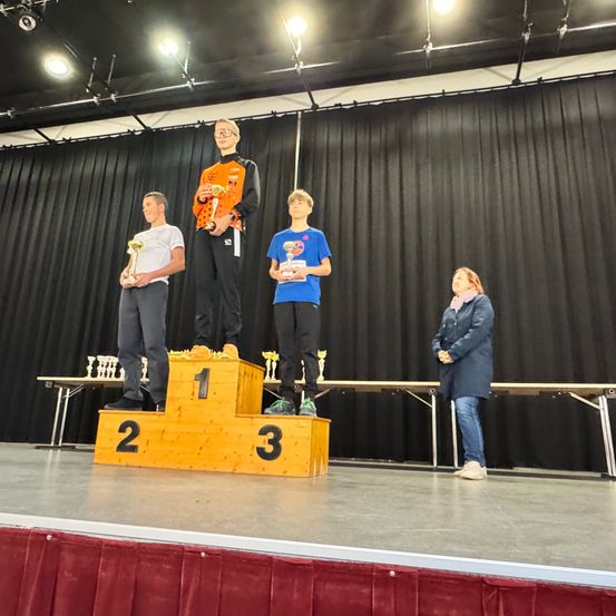 Drei Personen stehen auf einem Podium, eine oben und zwei unten. Sie halten alle Trophäen. Dahinter ist ein schwarzer Vorhang mit einem Tisch und Stühlen.