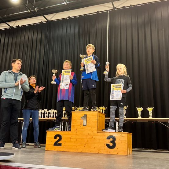 Drei Kinder stehen auf einem Podium und halten Pokale in den Händen. Sie sind von Erwachsenen umgeben. Das Podium ist aus Holz und hat die Zahlen 1, 2 und 3 darauf.
