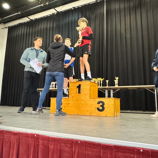 Ein Junge steht auf dem dritten Podestplatz und hält einen Pokal, während eine Person sein Bein hält. Hinter ihm hält ein Mann ein Papier und eine Frau steht neben ihm. Ein Junge in einem blauen Trikot steht links.