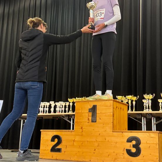 Eine Frau auf dem Podest hält einen Pokal und wird von einer anderen Frau unterstützt. Dahinter sind zahlreiche Pokale auf einem Tisch ausgestellt. Der Hintergrund ist ein schwarzer Vorhang.