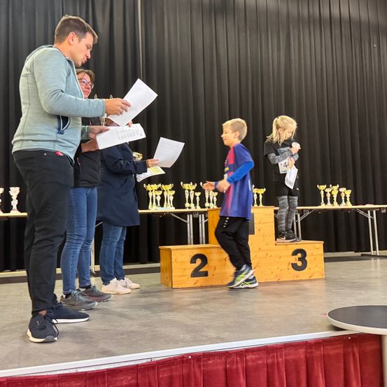 Auf der Bühne überreicht ein Mann einem Jungen ein Papier, der auf einem Podium steht. Dahinter hält ein weiteres Kind eine Trophäe. Zuschauer beobachten. Im Hintergrund stehen Trophäen und ein schwarzer Vorhang.