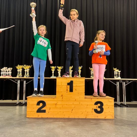 Drei junge Mädchen stehen auf einem Podium und halten Trophäen. Sie tragen farbenfrohe Langarmshirts und Hosen. Das Podium hat die Zahlen 1, 2 und 3 darauf. Hinter ihnen stehen Trophäen und ein schwarzer Vorhang.