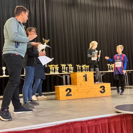 Ein Mann auf der Bühne überreicht zwei Kindern Pokale, eines hält einen Pokal und das andere lächelt. Dahinter sind Pokale auf einer Holzplattform ausgestellt.