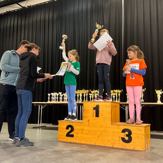 Drei junge Mädchen stehen auf einem Podium und halten Pokale. Sie lächeln und ein Mann und eine Frau, wahrscheinlich ihre Trainer, überreichen ihnen Urkunden. Das Podium hat die Zahlen 1, 2 und 3. Die Szene spielt sich in einem Raum mit schwarzen Vorhängen ab.