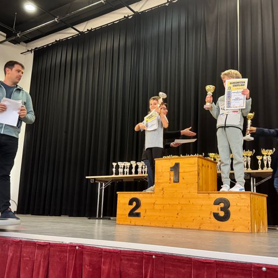 Eine Bühne mit Podestplätzen, nummeriert 1, 2 und 3. Ein Junge auf dem ersten Podest hält einen Pokal. Ein Junge auf dem zweiten Podest hält zwei Pokale. Ein Mann steht links.