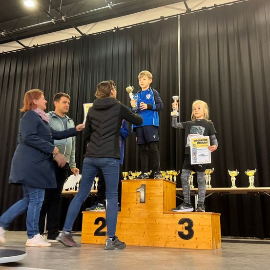 Drei Kinder stehen auf einem Podium in einer Turnhalle und halten Trophäen. Eine Frau überreicht dem Kind auf der rechten Seite eine Urkunde. Dahinter stehen Erwachsene in der Nähe eines Tisches mit Trophäen. Die Bühne ist vor schwarzen Vorhängen aufgebaut.