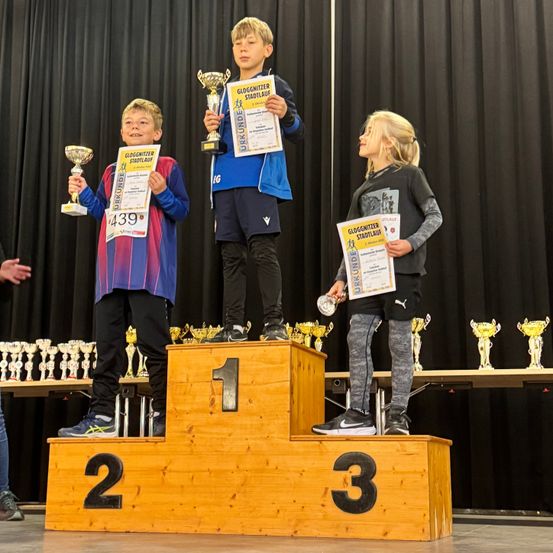 Drei Kinder stehen auf einem Podest, zwei halten Pokale, das dritte schaut zu. Sie tragen Sportbekleidung und halten Urkunden. Die Zahlen 1, 2 und 3 sind auf dem Podest zu sehen. Hinter ihnen befinden sich Pokale und ein Hintergrund.