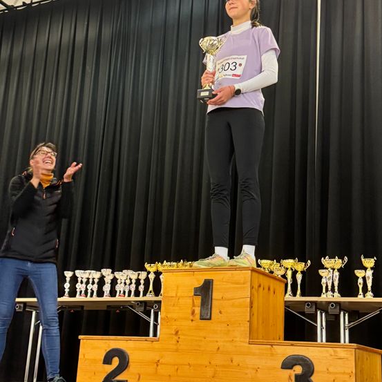 Eine Frau steht auf dem Siegerpodest und hält eine Trophäe, während eine andere Frau applaudiert. Sie stehen vor einem schwarzen Vorhang mit zusätzlichen Trophäen auf dem Podium und den Tischen.