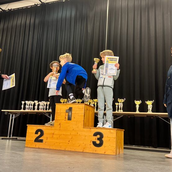 Drei Kinder stehen auf einem Podium mit den Nummern 1, 2 und 3 und halten Pokale. Sie tragen Sportbekleidung und haben Urkunden in den Händen. Hinter ihnen stehen schwarze Vorhänge und Tische mit Pokalen.