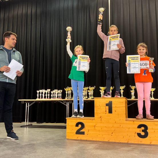 Drei junge Mädchen stehen mit Trophäen auf einem Podest und lächeln für die Kamera. Sie werden von einem Mann begleitet, der ein Stück Papier in der Hand hält. Dahinter sind verschiedene Trophäen ausgestellt.