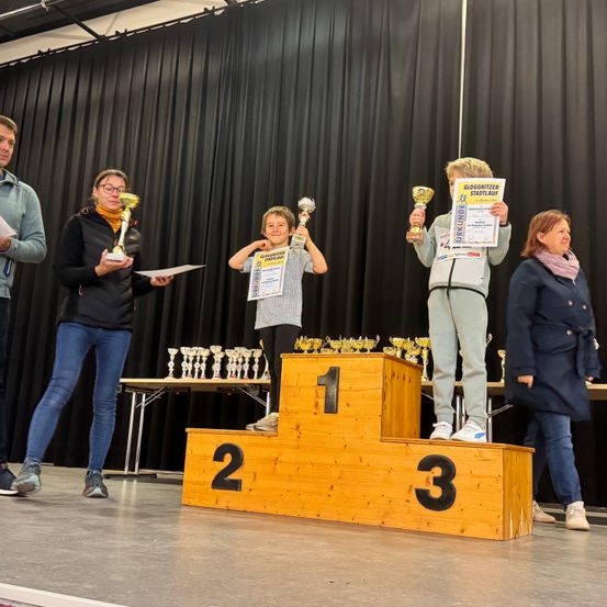 Eine Gruppe von Kindern und Erwachsenen auf der Bühne mit Pokalen. Die Bühne hat die Zahlen 1, 2 und 3 darauf. Dahinter steht ein Tisch mit weiteren Pokalen. Die Kinder lächeln und halten Urkunden in der Hand.