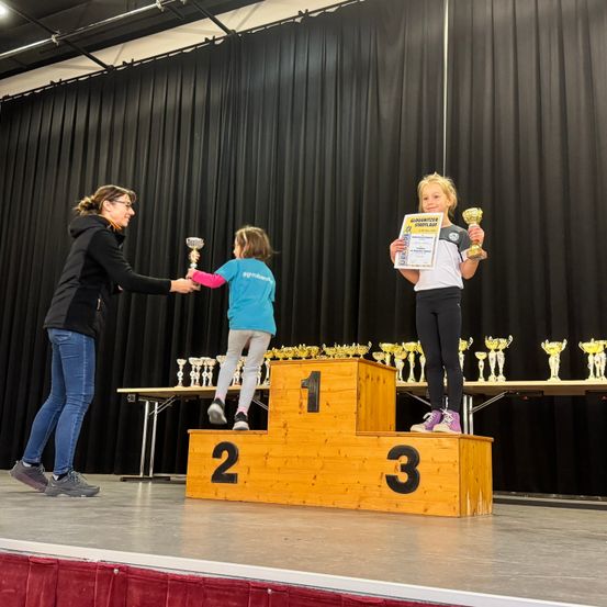 Ein junges Mädchen auf der Bühne erhält eine Trophäe von einer Frau, ein weiteres Kind in der Nähe, beide halten Trophäen, und ein Hintergrund aus schwarzen Vorhängen.